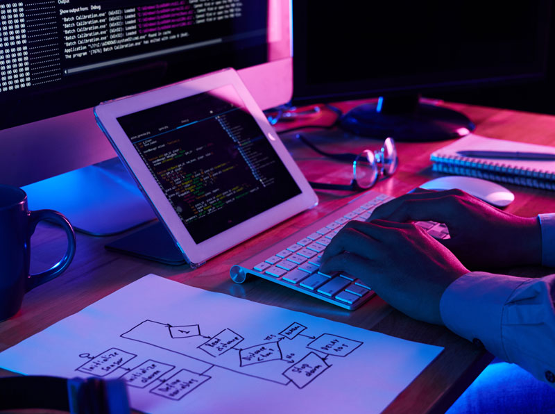 برنامه نویس با استفاده از یک زبان برنامه نویسی در حال کدزدن است و در کنار لب تاب و تبلت، یک کاغذ فلوچارت قرار دارد